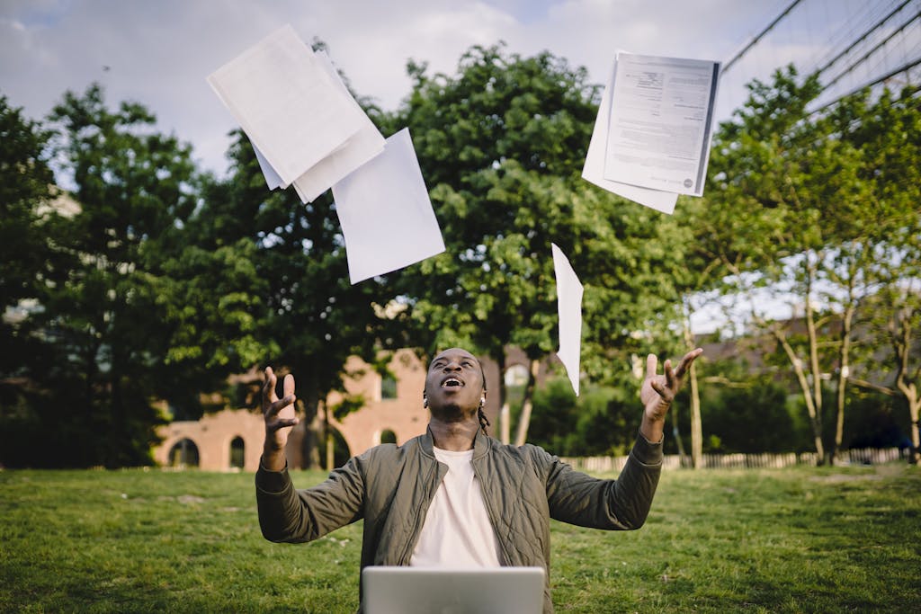 Overjoyed African American graduate tossing copies of resumes in air after learning news about successfully getting job while sitting in green park with laptop