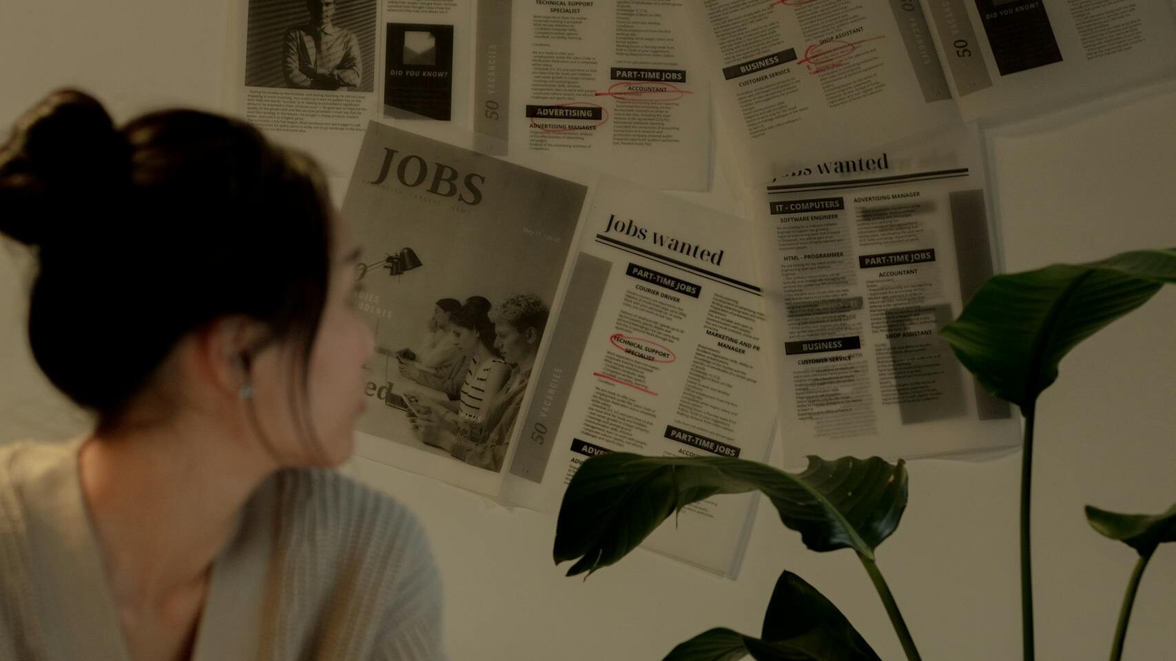 A Woman in Beige Sweater Sitting while Looking at the Newspapers on the Wall - Checking Job Market
