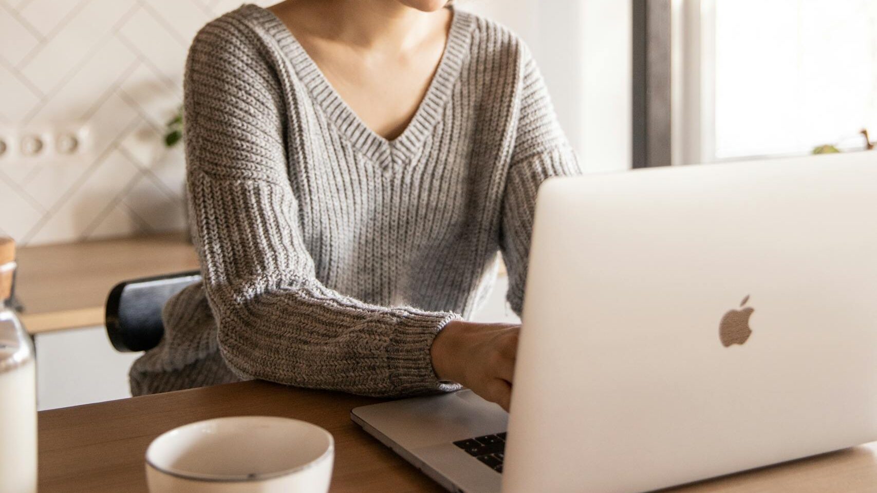 Young female in gray sweater sitting at wooden desk with laptop and bottle of milk near white bowl while browsing internet on laptop during free time at home, preparing for questions and learning advanced interview techniques
