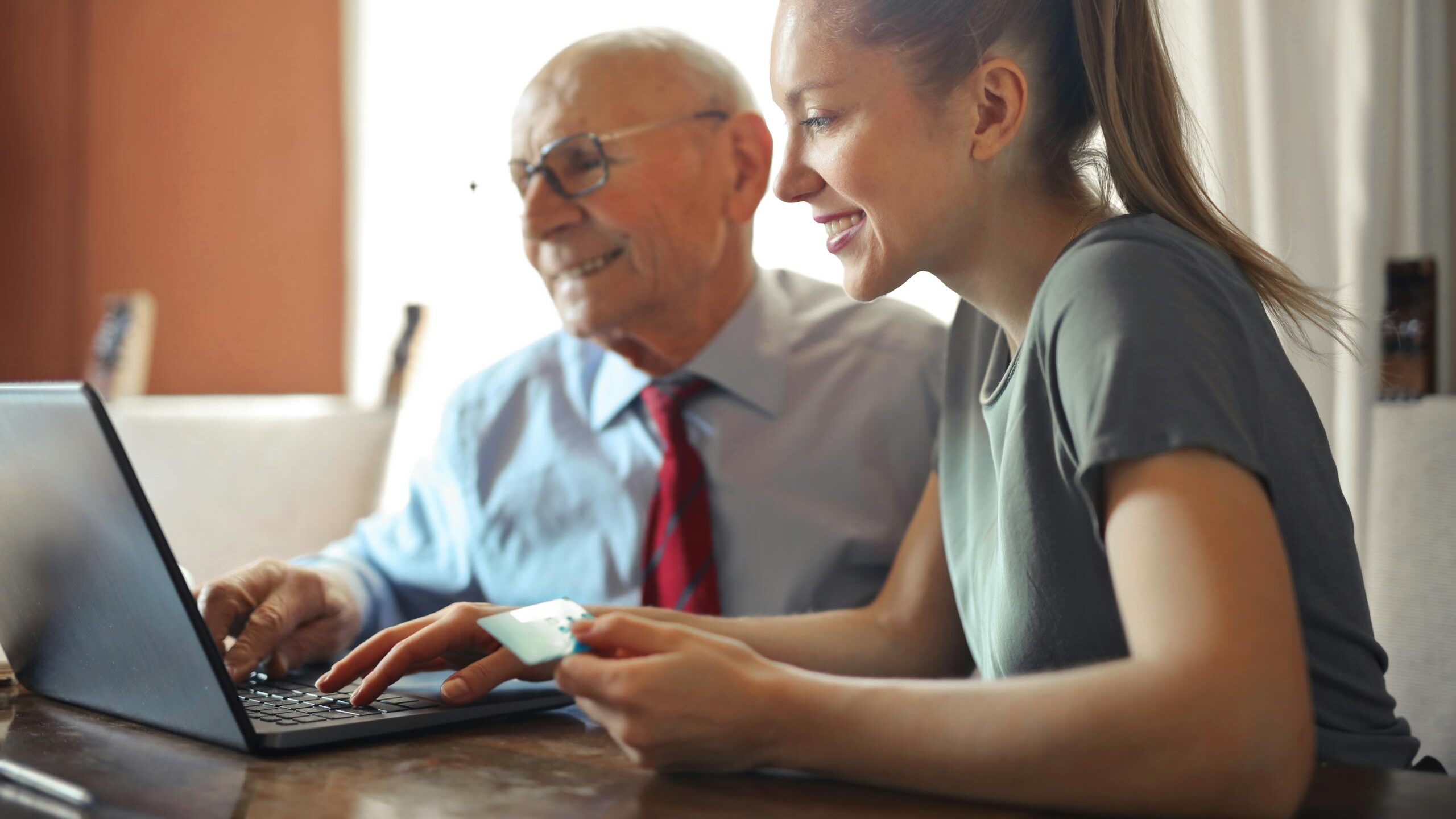 Young woman in casual clothes helping senior man in formal shirt with paying credit card in Internet using laptop while sitting at table