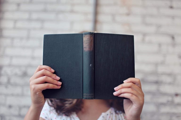 A woman holds an open book, reading in front of a brick wall.